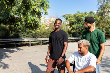 Young disabled Black man in a wheelchair and his male friends walking together in the city. Copy...