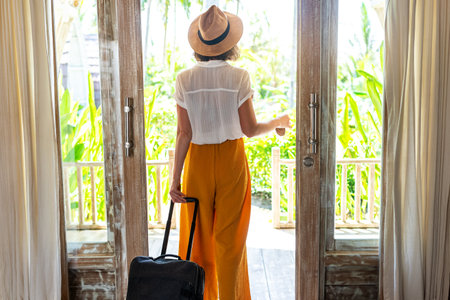 Rear view of unrecognizable woman tourist with hat leaving hotel bedroom with suitcase.の写真素材