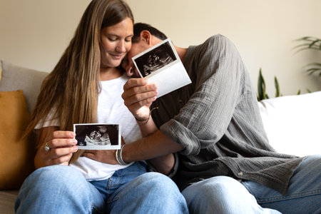 Happy and excited pregnant couple looking baby ultrasounds at home sitting on the couch. Man touching wife belly.の写真素材