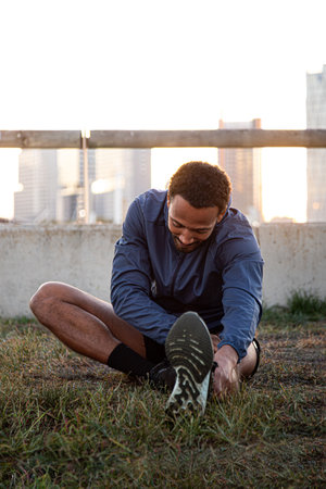 Vertical portrait of young african american man stretching leg before running, training outdoors.の写真素材