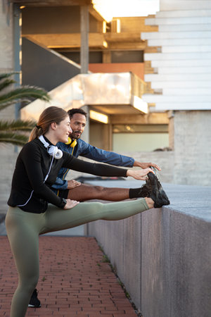 Vertical portrait of young multiracial couple stretching leg before running, training outdoors.