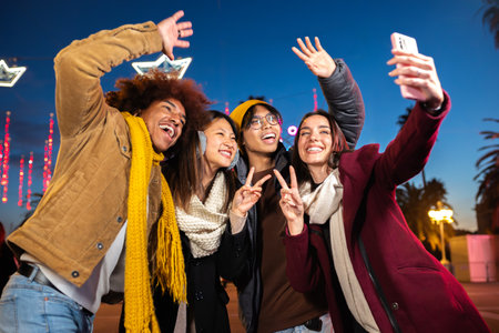Happy friends taking selfie at night in the city using phone. Friends having fun in Christmas winter market outdoors.の写真素材
