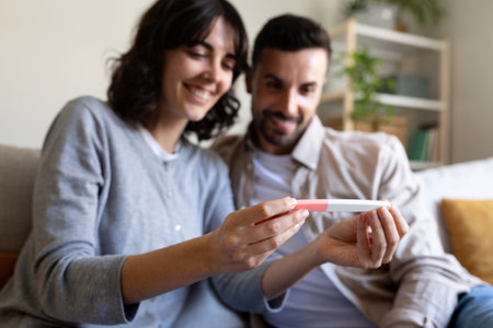 Happy caucasian couple looking at positive pregnancy test together at home. Selective focus on hands. Blurred backgroundの写真素材