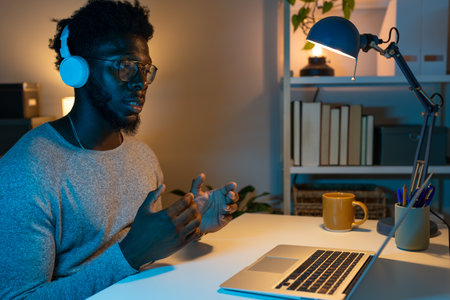 Black man working late at night at home office using laptop and headphones on a video call.