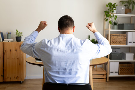 Back view of businessman celebrating success with arms raised up sitting at home office.の写真素材