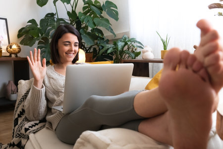 Young happy woman waving hand during online video call using laptop at home cozy living roomの写真素材