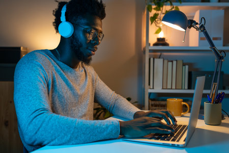 Black man working at home office using laptop and headphones. African American male student studying late at night.の写真素材