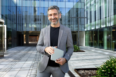 Happy mature man wearing a suit looking at camera outdoors in front of corporate business holding digital tablet.の写真素材
