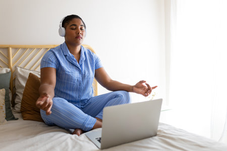 Serene Black woman meditating with headphones and laptop on bed in pajamasの写真素材