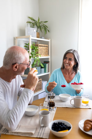 Vertical portrait of happy mature couple having healthy breakfast at home. Man eating orange, woman serving raspberries.の写真素材