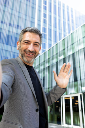 Vertical selfie of happy mature man in the city near office buildings looking at camera waving hand.の写真素材