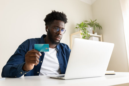 Young African American man online shopping using laptop and credit card at home office.の写真素材
