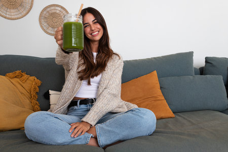 Happy woman showing healthy green juice looking at camera sitting on the couch. Female enjoying vegetable smoothie.の写真素材