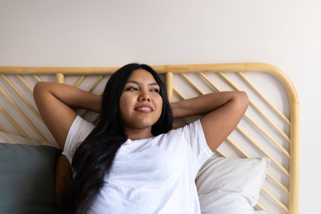 Young latin american woman relaxing in bed with hands behind headの写真素材