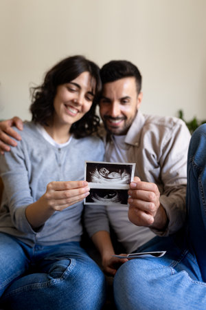 Vertical portrait of happy pregnant couple looking baby ultrasounds at home. Selective focus on hands.の写真素材