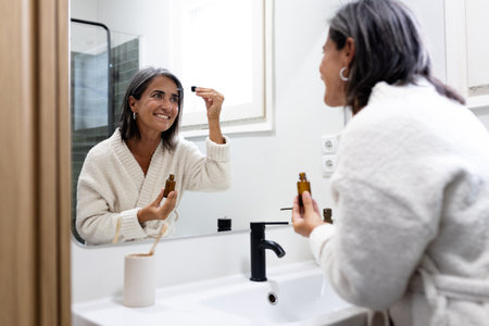 Mature woman gently applying face oil serum in bathroom.の写真素材