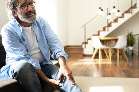 Mature man having knee pain sitting on the sofa at home living room. Copy space.の写真素材