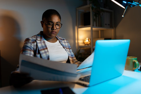 Young Black woman working late reviewing paperwork illuminated by laptop screenの写真素材