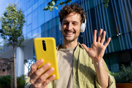 Young man wearing headphones is video calling with smartphone and waving in a modern cityの写真素材