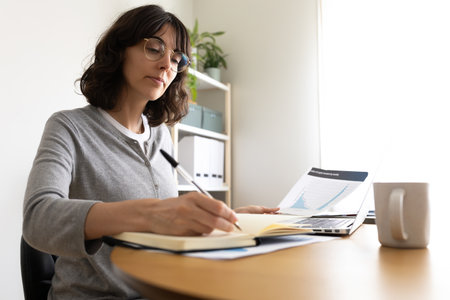 Woman writing on notebook, working at home office using laptopの写真素材
