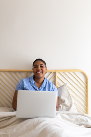 Happy black woman using laptop in bed working remotely from home looking at cameraの写真素材