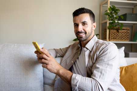Happy man using mobile phone at home relaxing on the sofa looking at camera smiling. Copy space.の写真素材