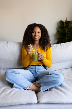 Vertical portrait of Black woman relaxing at home sitting on the sofa drinking green juice looking at camera.の写真素材