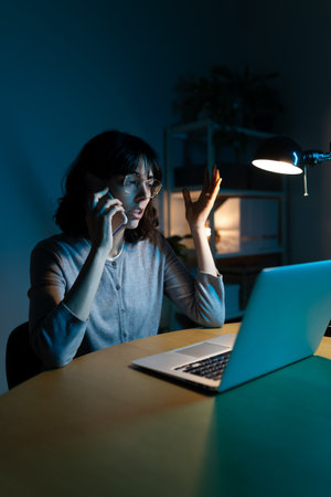 Stressed woman feeling overwhelmed talking on the phone while working at home office at night. Verticalの写真素材