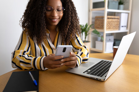 Happy African American woman using phone working at home office.の写真素材