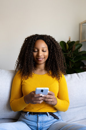 Vertical portrait of African American woman using phone relaxing at home living room sitting on couchの写真素材