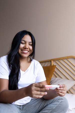 Happy latin american woman holding pregnancy test sitting on bedの写真素材