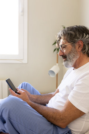 Side view vertical portrait of mature man using phone relaxing on bed at home.の写真素材