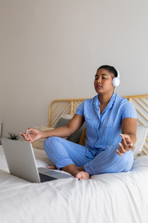Young African American woman meditating with headphones and laptop on bed in pajamasの写真素材