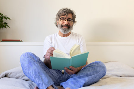 Mature man relaxing in bed, reading a book wearing pajamas looking at camera.の写真素材
