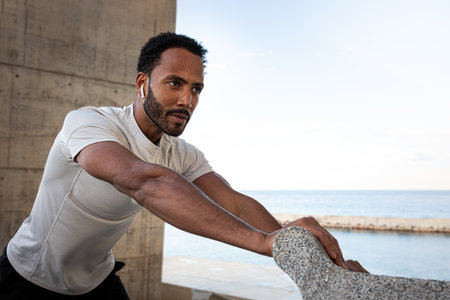 Focused athlete stretching by the sea, listening to music on wireless earbuds