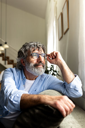 Pensive mature man with glasses relaxing sitting on the sofa looking out the window. Vertical imageの写真素材