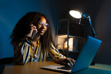 Happy Black woman talking on the phone while working at home office at night using laptop.の写真素材