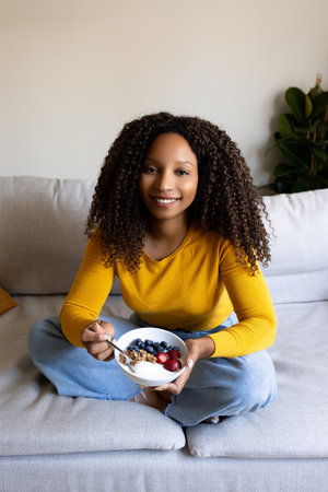 Black woman eating healthy breakfast sitting on the couch at home cozy living room looking at camera. Verticalの写真素材