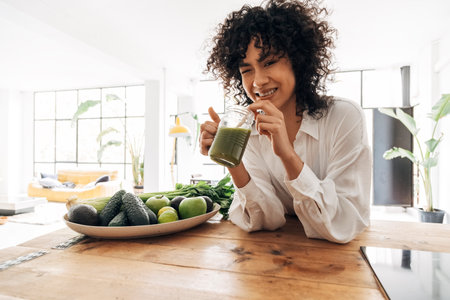 Young african mixed race woman looking at camera drinking green juice with bamboo straw in big loft kitchenの写真素材