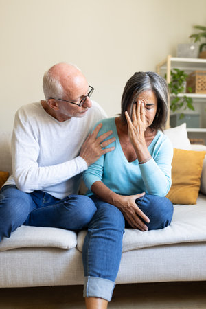 Vertical portrait of mature man consoling sad wife sitting on the sofa at home living room.の写真素材