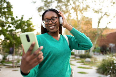Happy young black woman wearing headphones and using phone in a college campusの写真素材
