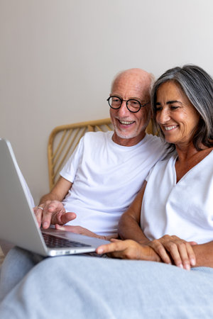 Vertical portrait of mature happy heterosexual couple using laptop together relaxing sitting on the bed.の写真素材