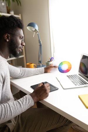 Black male graphic designer working at home using laptop and graphic tablet. Vertical imageの写真素材