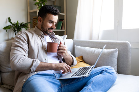 Adult man typing on laptop, sitting on the sofa drinking coffee. Male working from home.の写真素材
