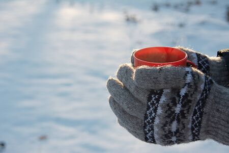 mug with hot drink in hands dressed in knitted wool mittens in winter on a Sunny frosty day, winter...
