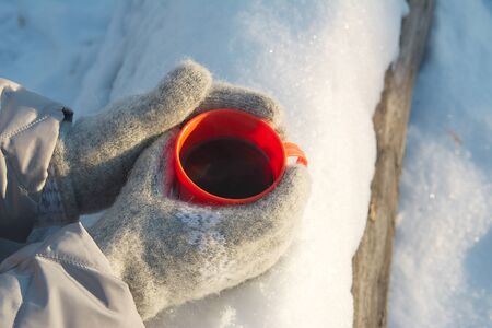 mug with hot drink in hands dressed in knitted wool mittens in winter on a Sunny frosty day, winter picnic, winter moodの写真素材