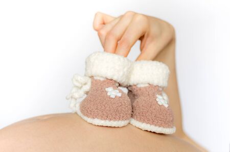 female hand holding knitted booties for newborn babies on background of the pregnant belly, the...