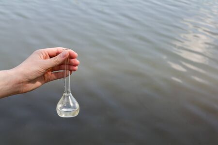 hand holding test tube for analyses with water on the background of the reservoir, the concept of water purity, pollution of water bodies, checking the quality of drinking water in cities, environmentalの写真素材