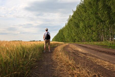 young man traveler with backpack in hat stands in field at sunset and looks away, concept of freedom of choice, future, concept of travel and adventureの写真素材
