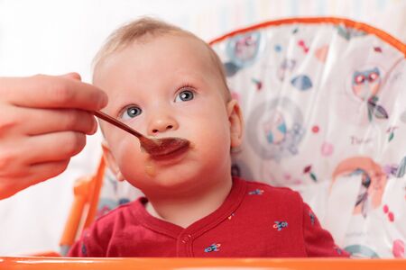 a small child eats with a spoon and sits and holds the edge of the feeding table in the nursery, grimy and stained with food, baby food concept, complementary foods, the first spoonの写真素材